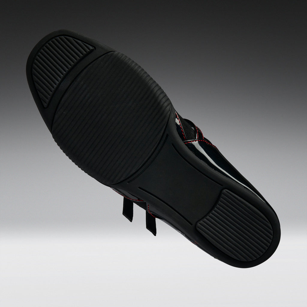 A black athletic shoe shown from its sole side, featuring a textured black rubber outsole and thin red contrast stitching on the upper, set against a dark gray gradient background.