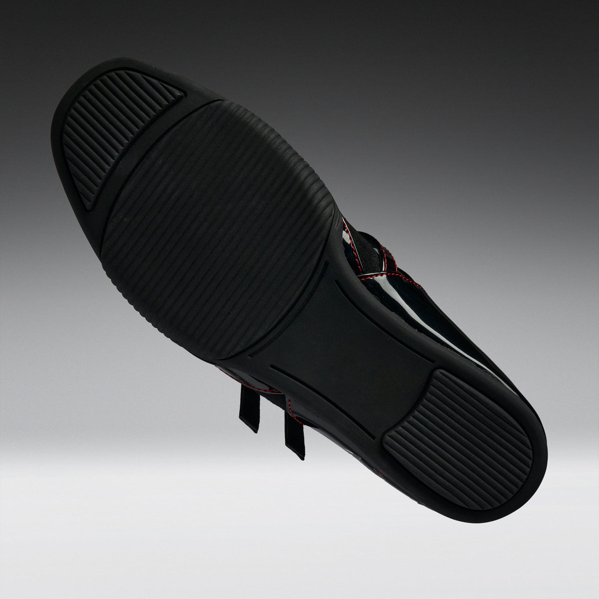 A black athletic shoe shown from its sole side, featuring a textured black rubber outsole and thin red contrast stitching on the upper, set against a dark gray gradient background.