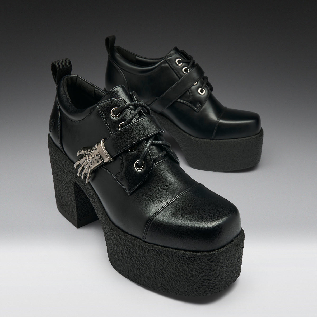 A pair of black leather platform lace-up shoes with chunky block heels, decorative straps, and silver hardware, photographed on a dark-to-light gray gradient studio background.