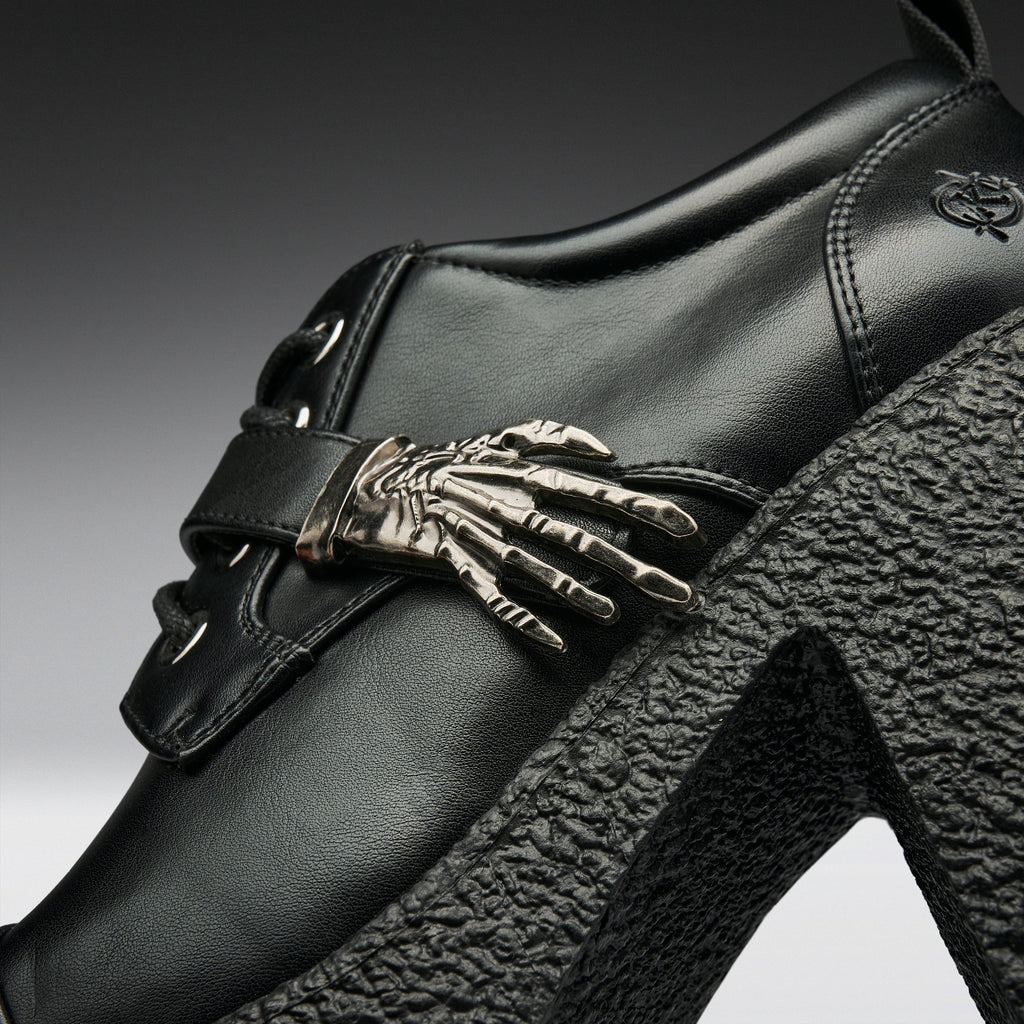 Close-up of a black leather high-heeled shoe featuring a prominent metallic skeleton hand ornament on the side strap, a chunky black textured platform heel, and a small embroidered logo near the heel.