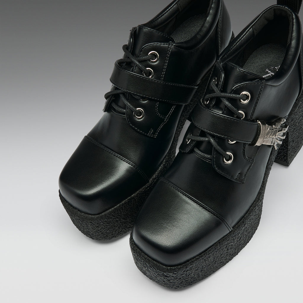 A pair of black platform lace-up ankle boots with chunky black soles, silver eyelets, and a small silver decorative accent on the side, shown against a gray gradient background.