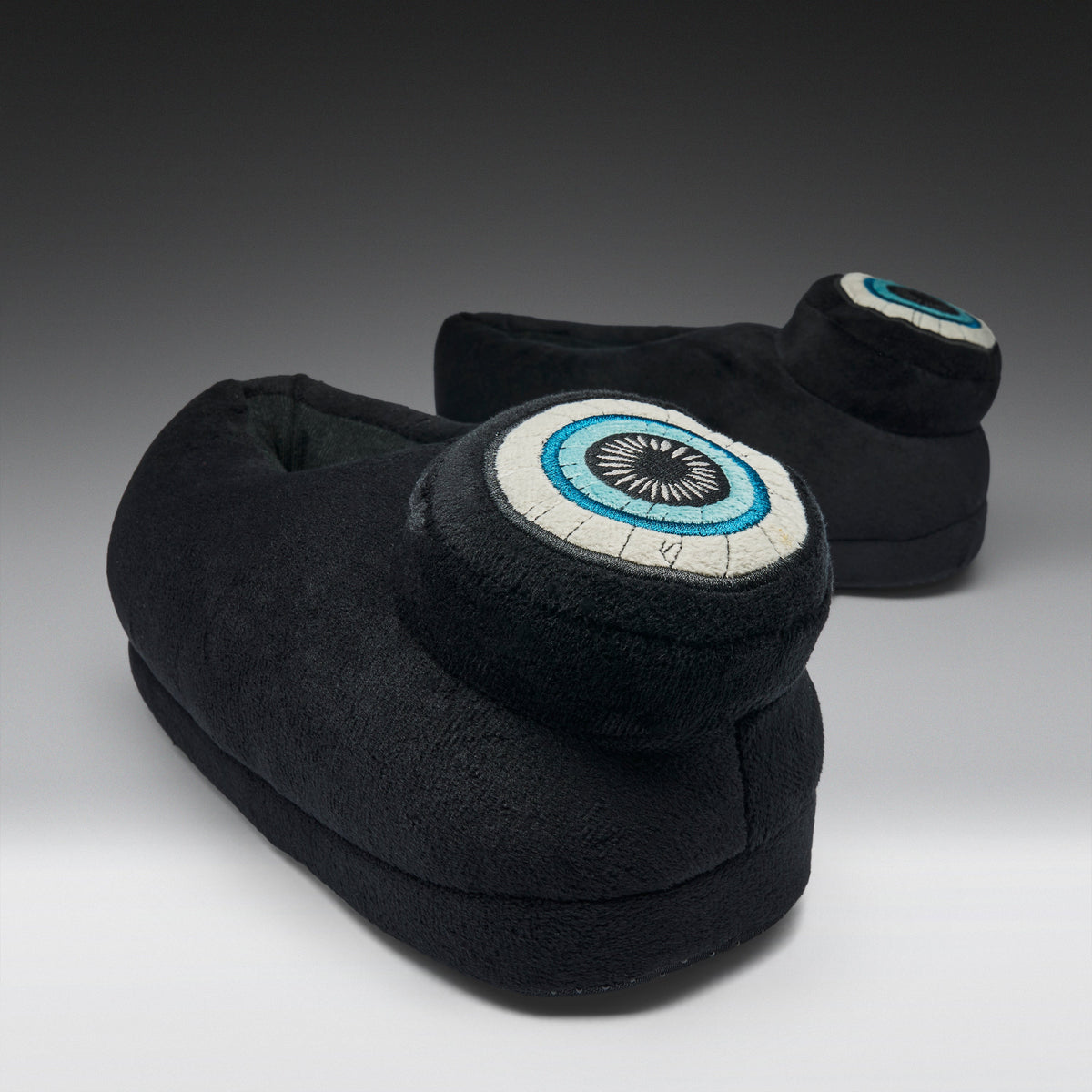 A pair of black plush slipper boots with circular eye motifs on the back, photographed on a dark-to-light gray gradient studio background.