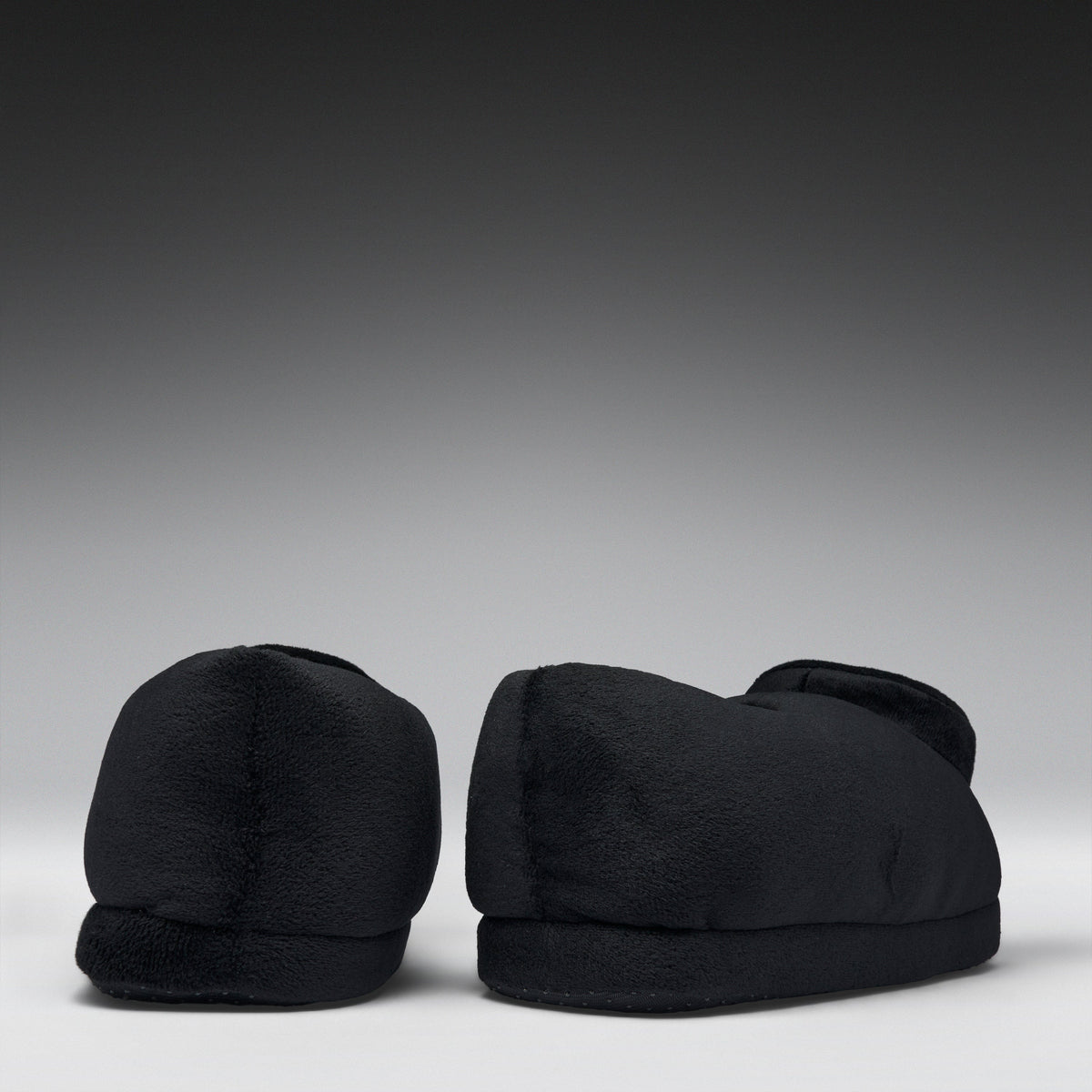 A pair of black plush slippers shown from the back against a dark gray gradient studio background.