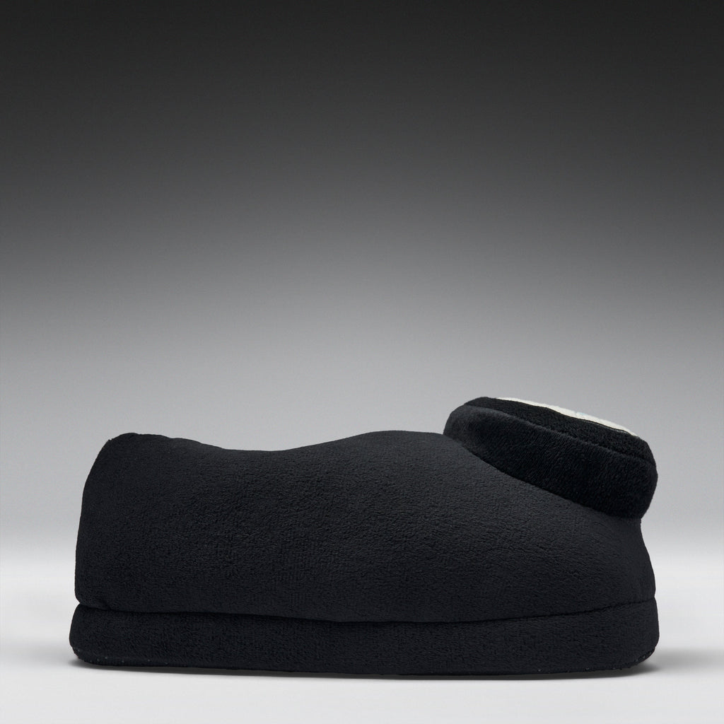 A black fleece-covered footrest or cushion with a curved shape and a small circular white accent on top, photographed against a dark gray to light gray gradient studio background.