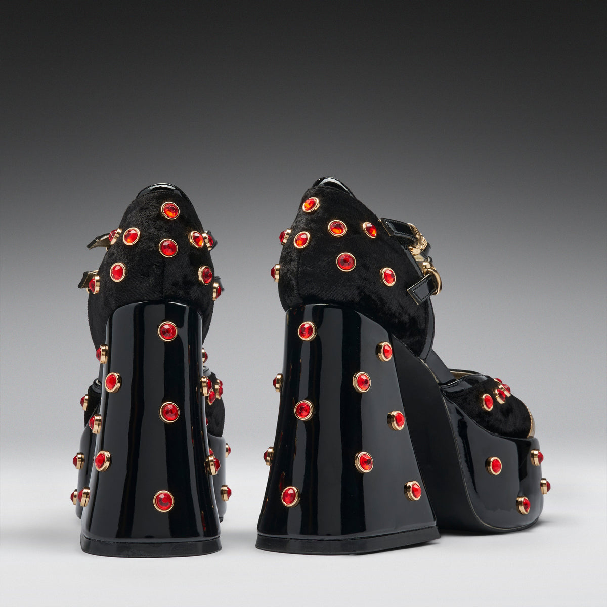 A pair of black platform high heels with glossy patent block heels, fuzzy black uppers, and numerous red rhinestone studs with gold-toned settings, shown from the back against a dark-to-light gray gradient studio background.