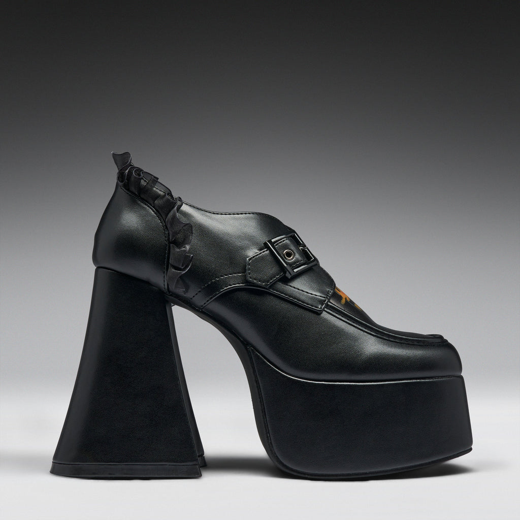 A single black platform high-heel shoe with a chunky sculptural block heel, a buckle strap across the instep, and a small orange accent near the toe, photographed against a dark-to-light gray gradient studio background.