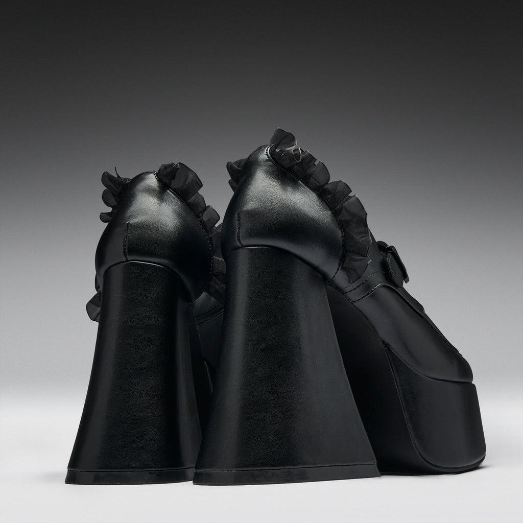A pair of black platform high heels with dramatic ruffled trim along the sides, photographed from the back against a dark gray gradient studio background.