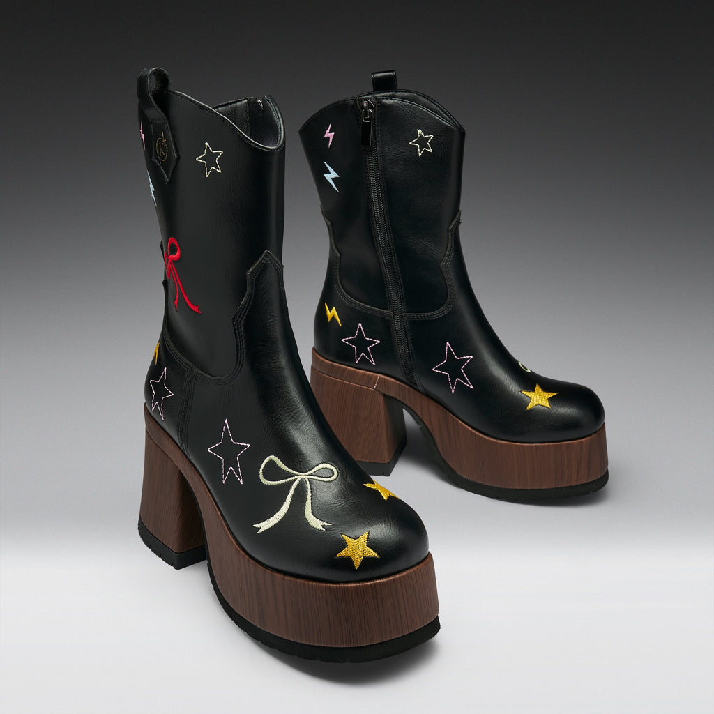 A pair of black leather ankle boots with brown platform heels, decorated with embroidered stars, bows, and lightning bolts in multiple colors, shown against a dark gray gradient background.