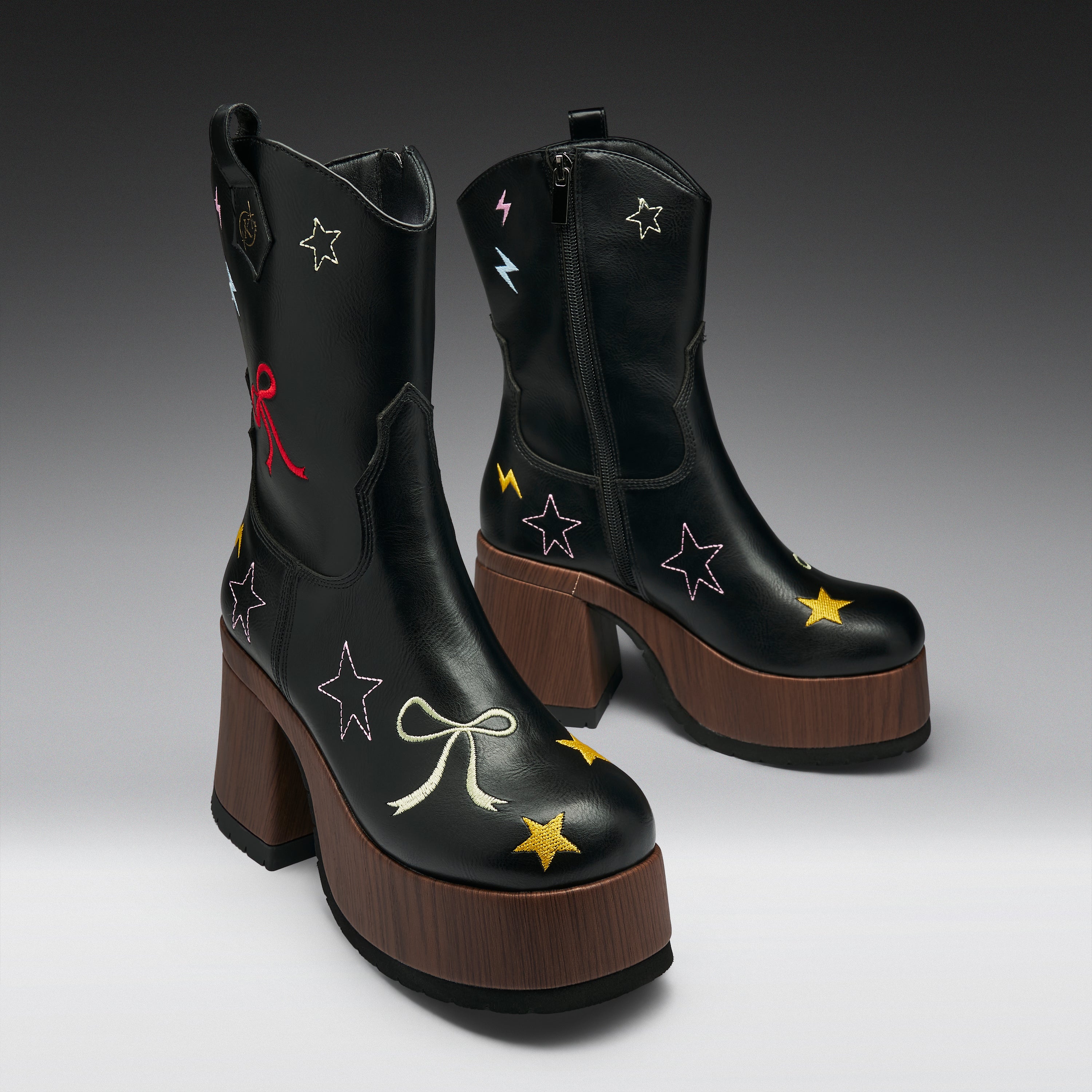 A pair of black leather ankle boots with brown platform heels, decorated with embroidered stars, bows, and lightning bolts in multiple colors, shown against a dark gray gradient background.