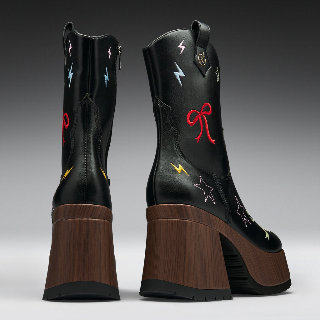 A pair of black leather mid-calf boots with chunky brown wooden platform heels, decorated with colorful embroidered stars, lightning bolts, and bows on a gray gradient studio background.