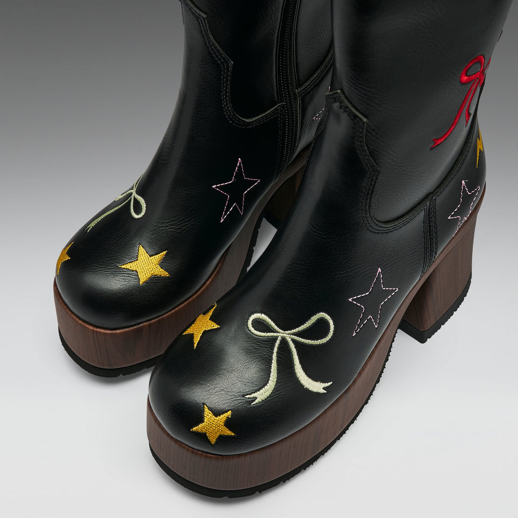 A pair of black leather western-style boots with brown platform soles, decorated with embroidered stars and bows in yellow, white, pink, and red on a light gray studio background.
