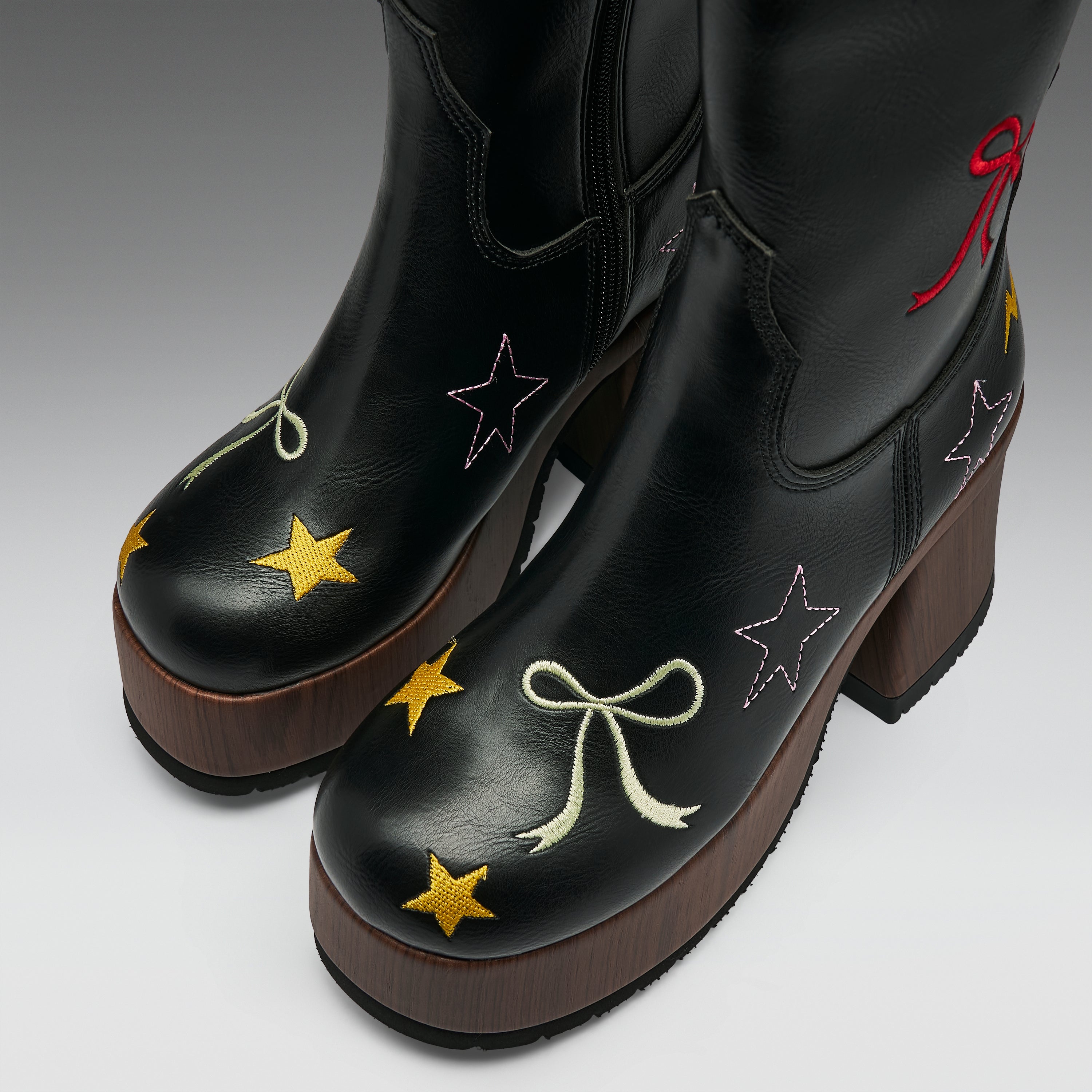 A pair of black leather western-style boots with brown platform soles, decorated with embroidered stars and bows in yellow, white, pink, and red on a light gray studio background.