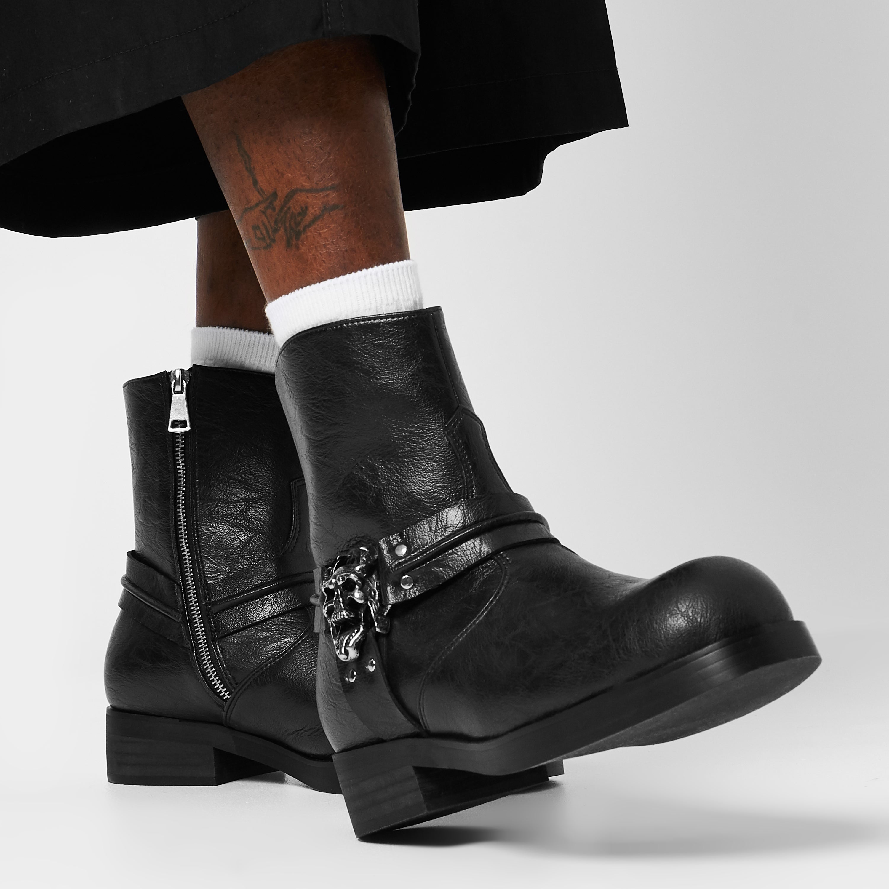 A close-up of a man's black biker boot with a stacked sole and hardware detail, worn with cropped trousers.