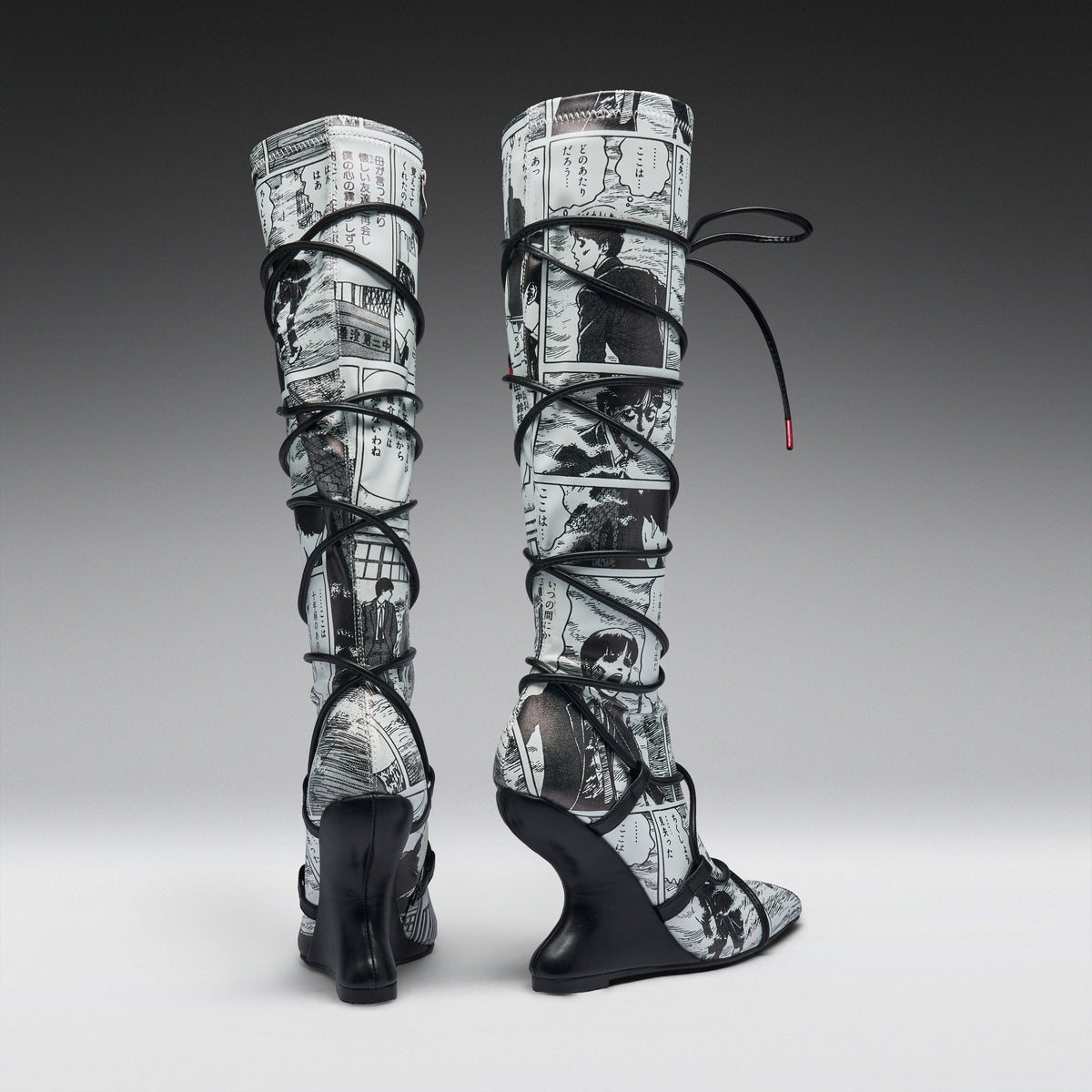 A pair of high-heeled, over-the-knee boots featuring a black and white newspaper print pattern wrapped around the shafts, with black lace-up straps and wedge heels, shown against a gray gradient studio background.