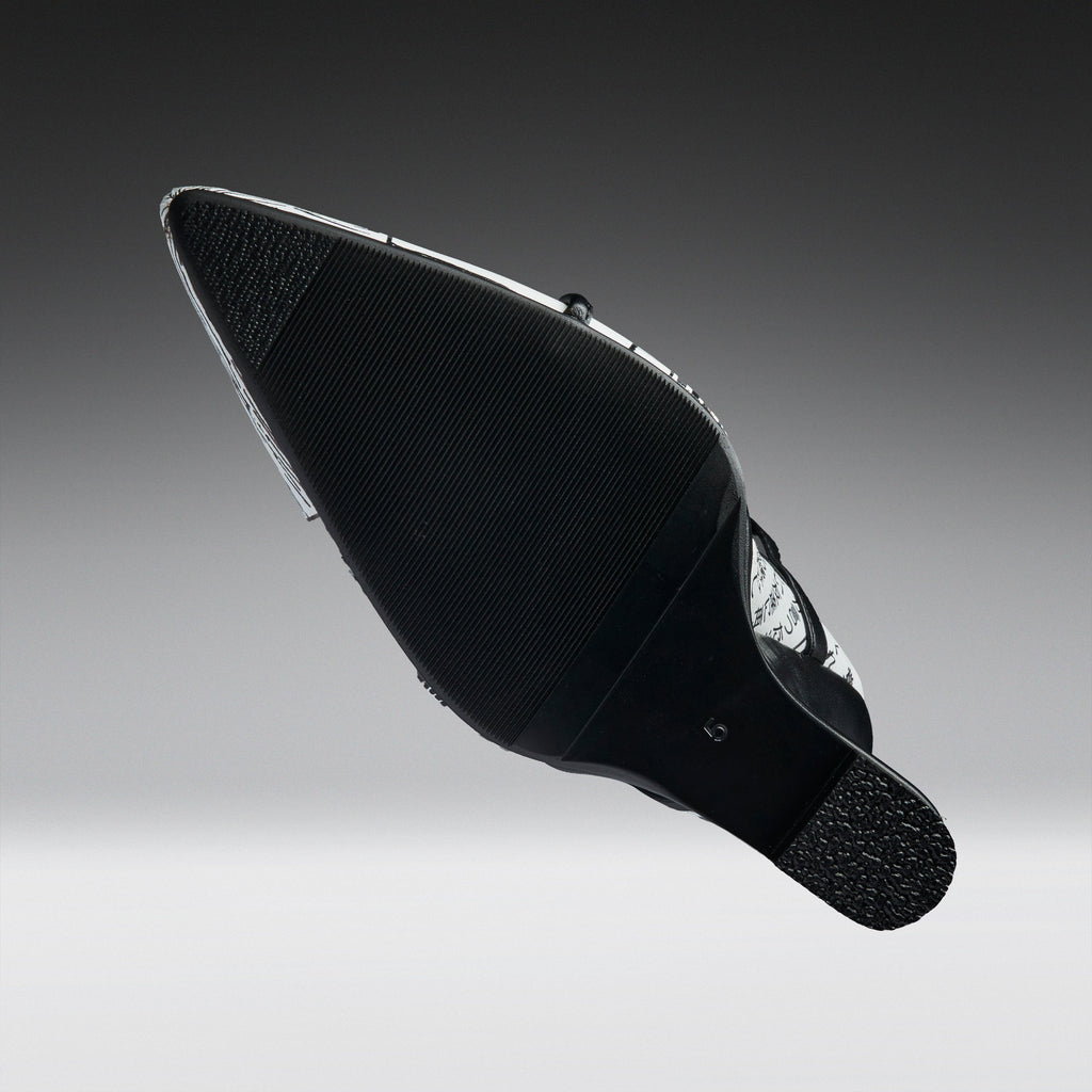 A black pointed-toe shoe shown from the sole side, featuring a textured black rubber outsole and a silver metallic side accent, set against a dark-to-light gray gradient background.