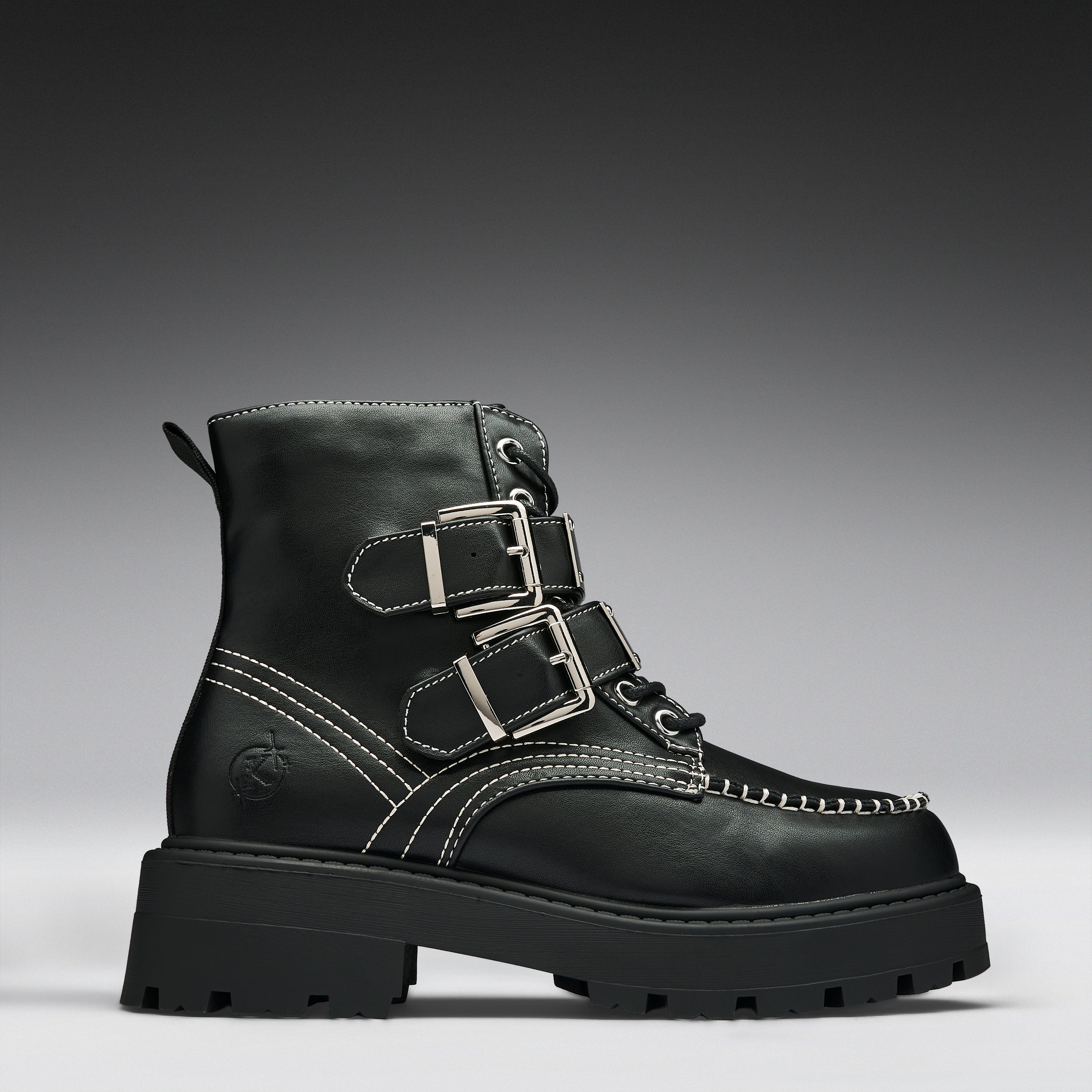 A black leather ankle boot with three silver buckle straps, white contrast stitching, and a chunky lug sole, shown against a dark gray gradient background.