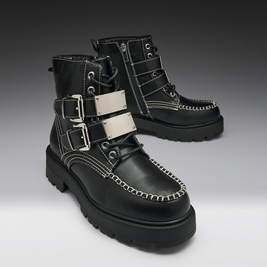 A pair of black leather lace-up ankle boots with silver buckle straps, white contrast stitching, and chunky black soles, shown on a dark-to-light gray gradient studio background.