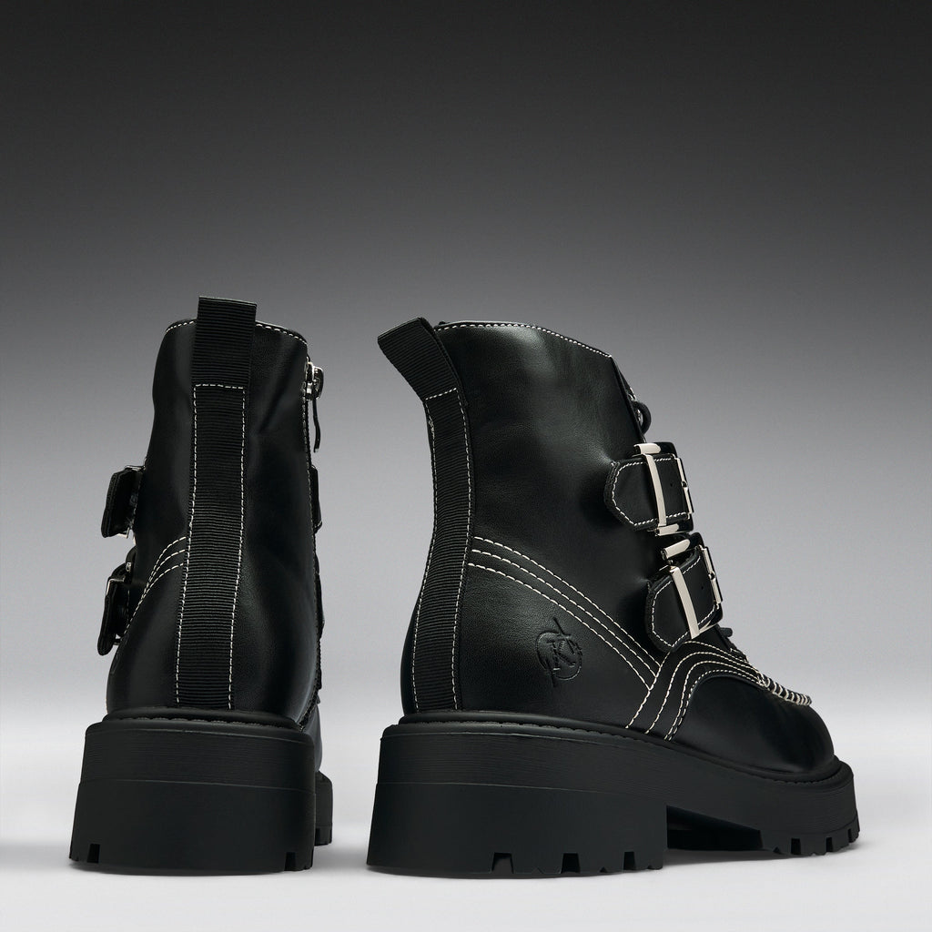 A pair of black leather ankle boots with chunky lug soles, featuring silver buckle details, contrast white stitching, and side zippers, shown from the back and side against a dark gray gradient background.