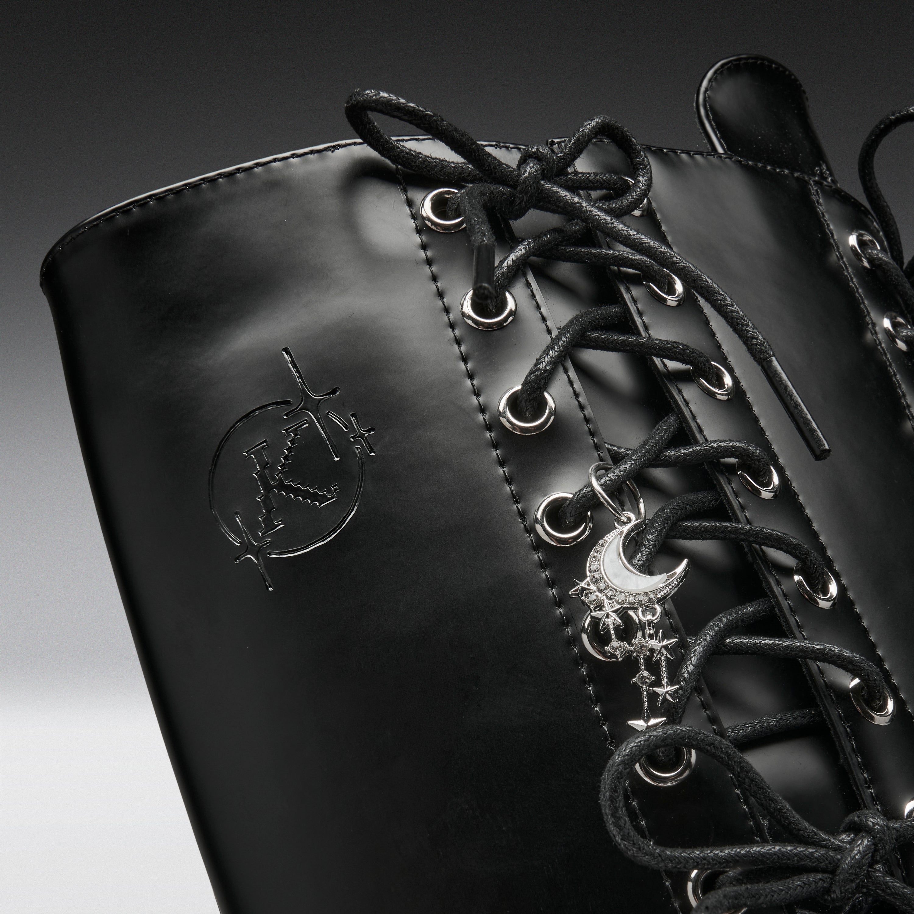 Close-up of a black leather lace-up boot with silver eyelets, a crescent moon and skull charm, and an embossed logo on the side.