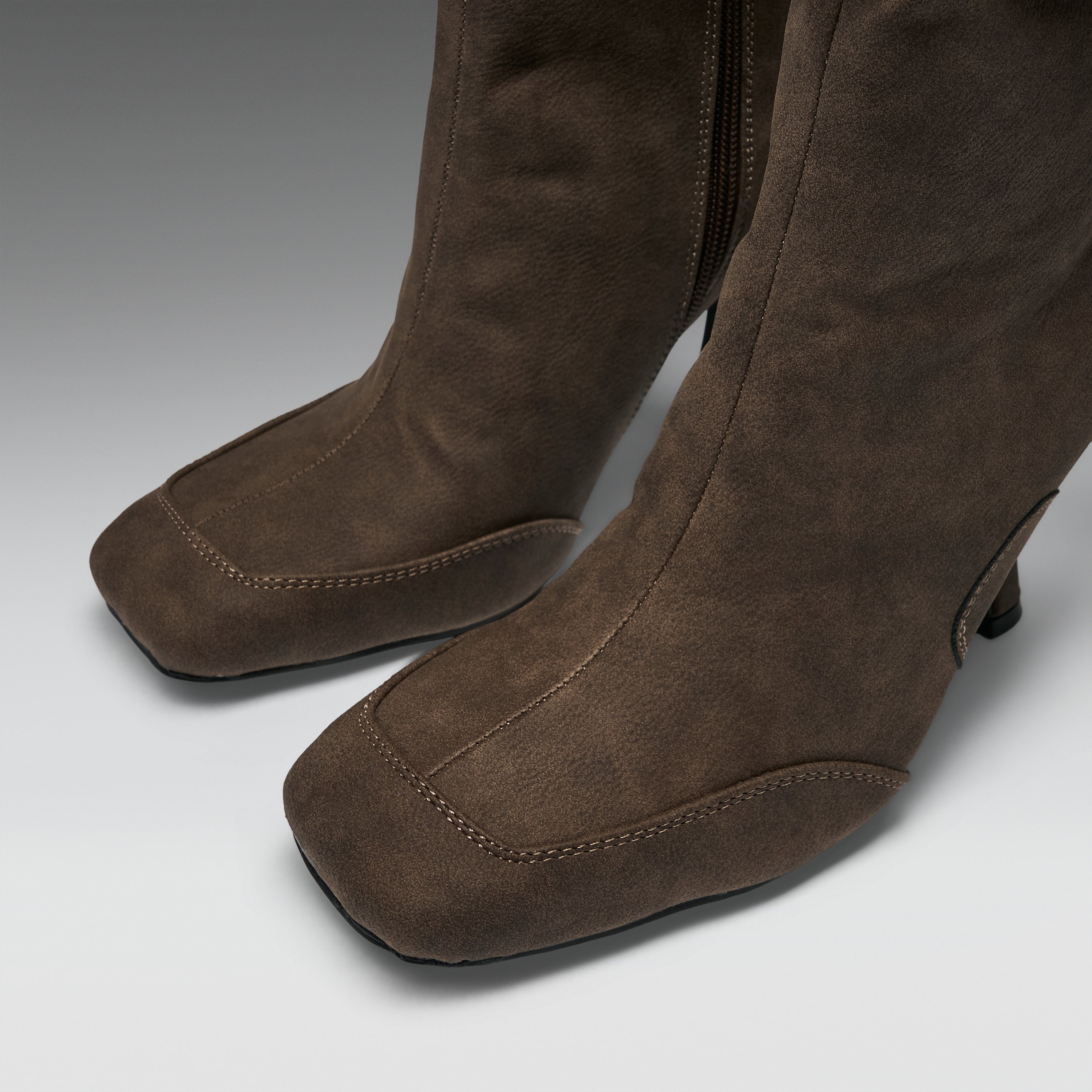 A close-up of a pair of brown suede over-the-knee boots with a square toe and low block heel, shown on a neutral gray background.