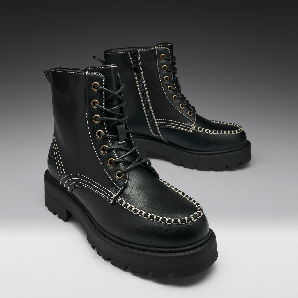 A pair of black leather lace-up boots with chunky lug soles, contrast white stitching, and brass eyelets, photographed on a dark-to-light gray gradient studio background.