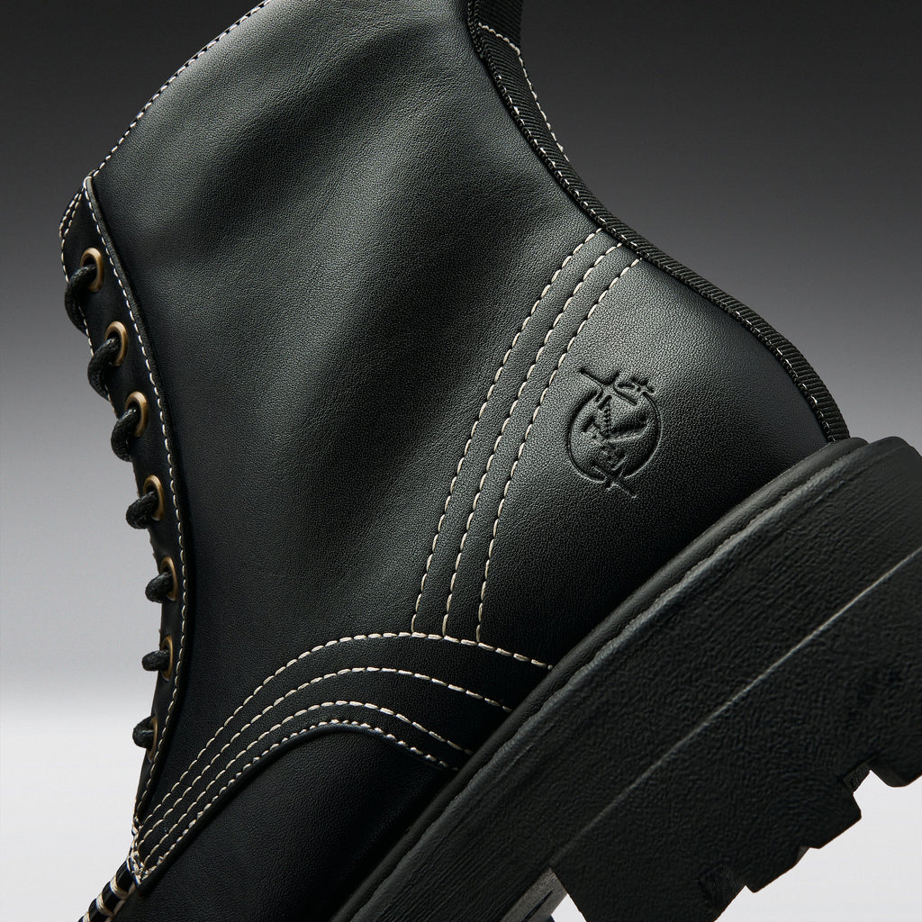 Close-up of a black leather lace-up boot with white contrast stitching, gold eyelets, and an embossed circular logo on the side, shown against a dark gray gradient background.