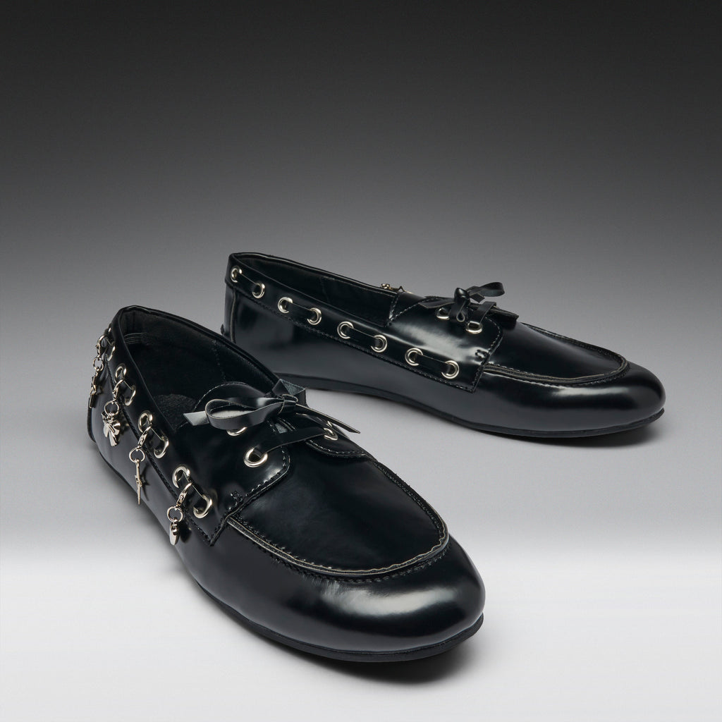 A pair of glossy black leather loafers decorated with silver-tone metal eyelets, tassels, and hanging charms, displayed on a dark-to-light gray gradient studio background.
