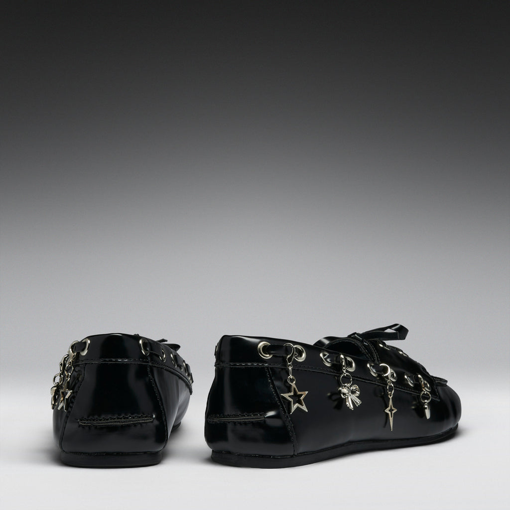 A pair of black patent leather lace-up flats decorated with silver-tone metal charms, including a star, cross, and clover, shown from the back and side against a dark gray gradient background.