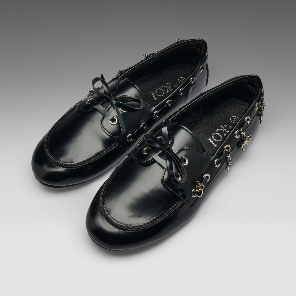 A pair of black leather boat-style loafers with decorative gold-tone hardware, bow ties, and dangling charms, displayed on a neutral gray gradient background. The brand name 'IOX' is visible on the insole.