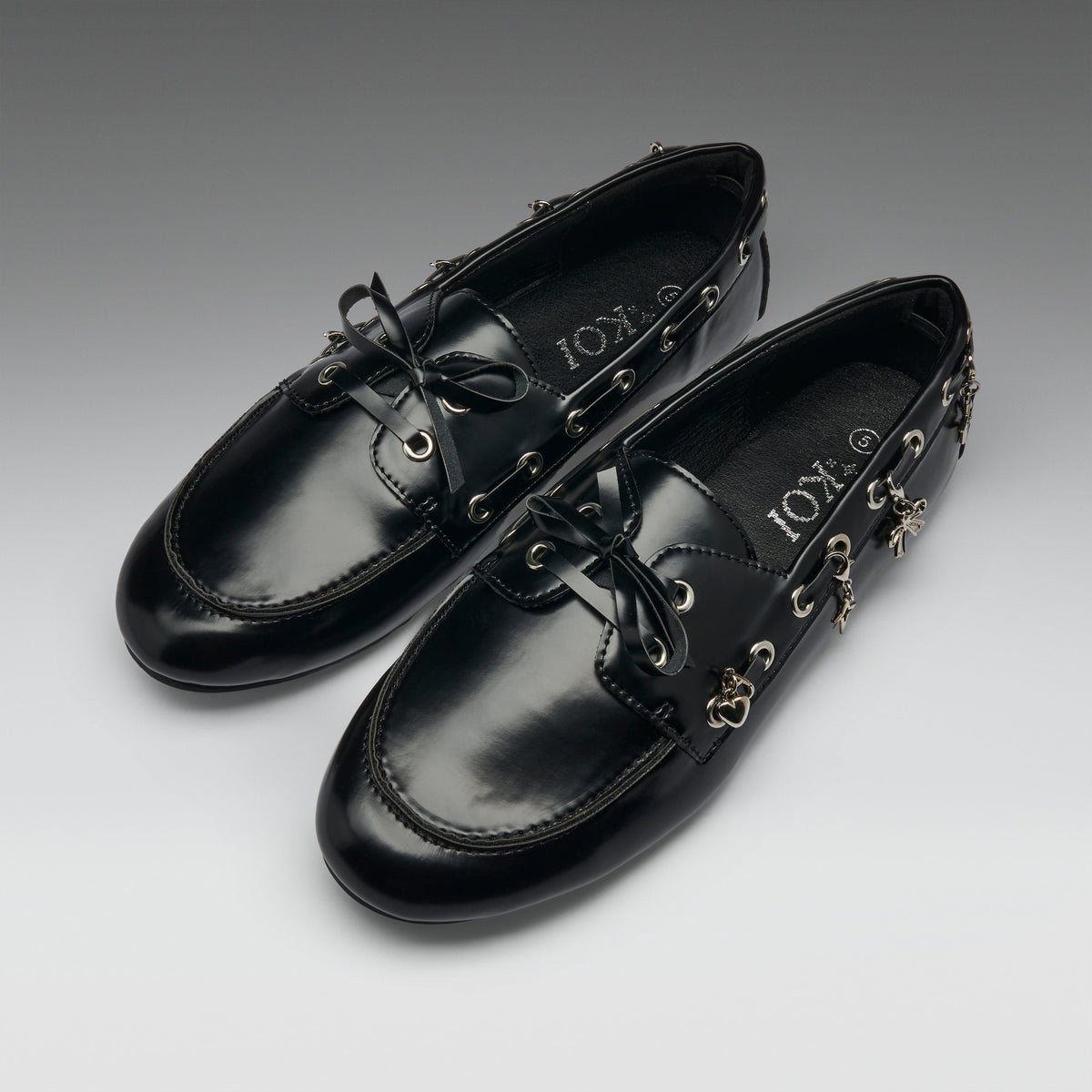 A pair of black leather boat-style loafers with decorative gold-tone hardware, bow ties, and dangling charms, displayed on a neutral gray gradient background. The brand name 'IOX' is visible on the insole.