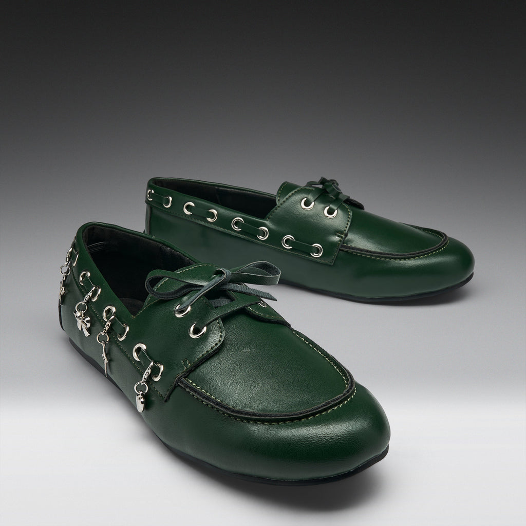 A pair of dark green leather boat-style shoes with decorative silver-tone metal eyelets, hanging charms, and matching green laces, displayed on a dark-to-light gray gradient studio background.
