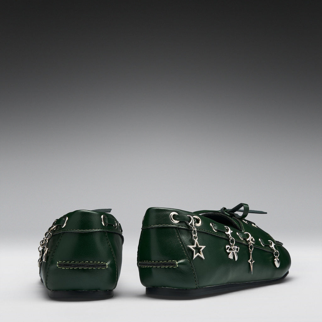 A pair of dark green leather baby booties decorated with silver-tone metal charms, including a star and bows, shown from the back on a gray gradient background.