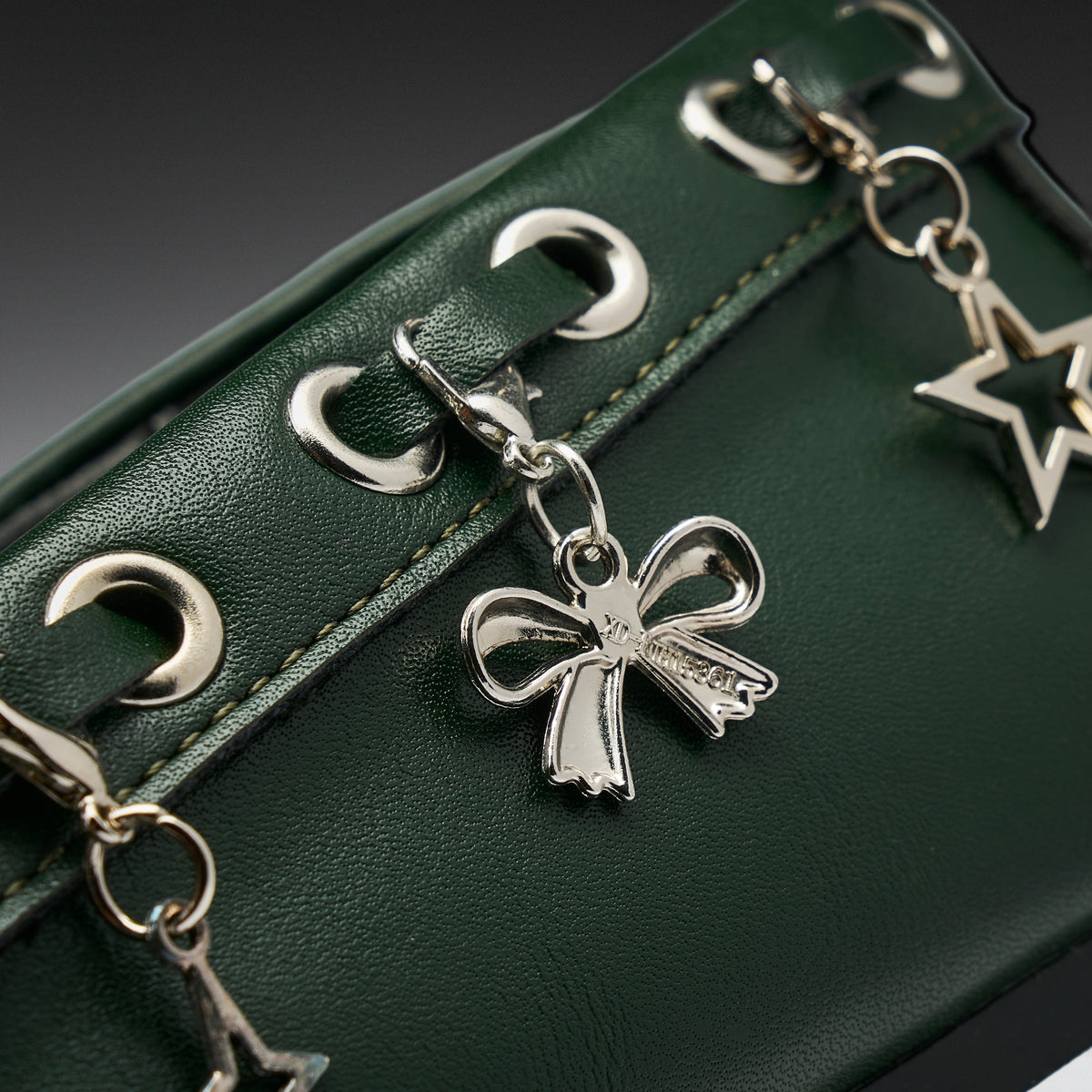 Close-up of a dark green textured leather bag featuring silver-tone metal grommets and dangling charms, including a bow-shaped charm and a star-shaped charm.