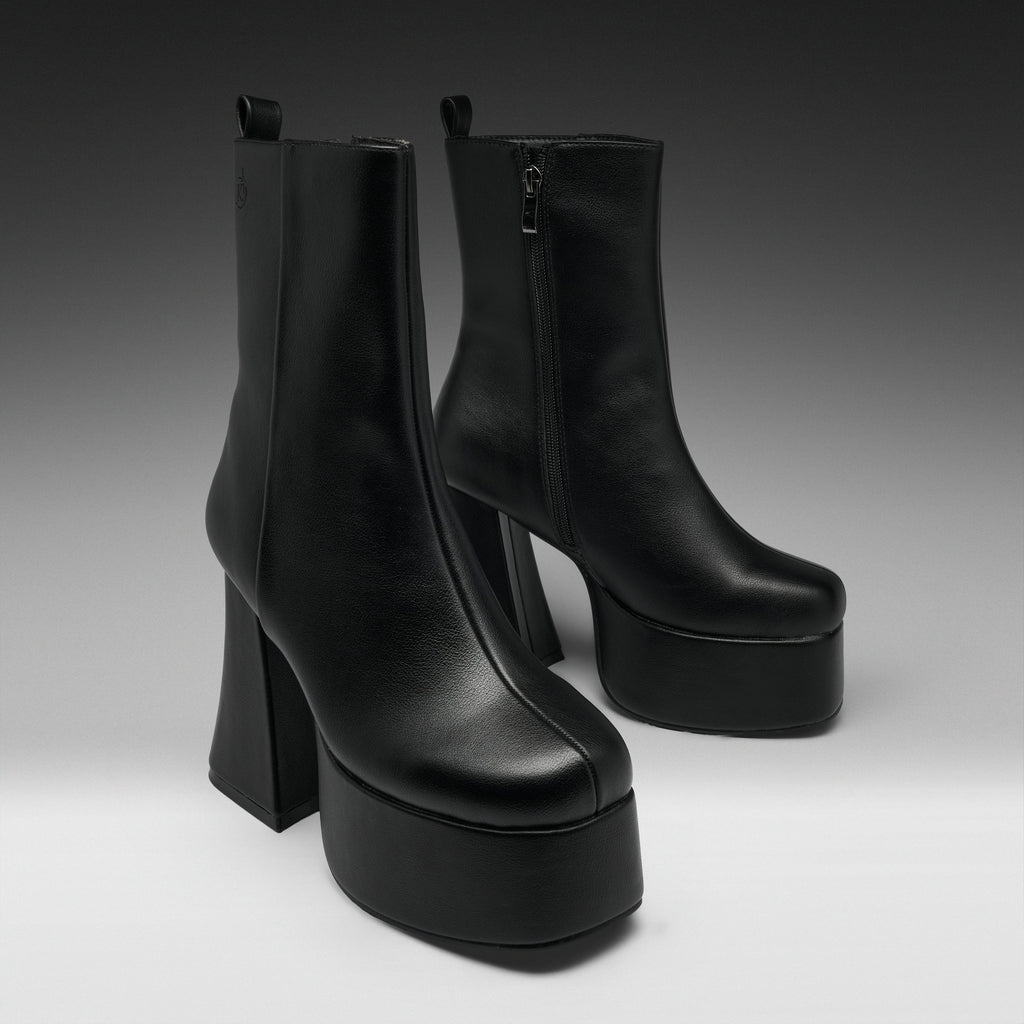 A pair of black leather ankle boots with high block heels and thick platform soles, photographed on a dark-to-light gray studio background.