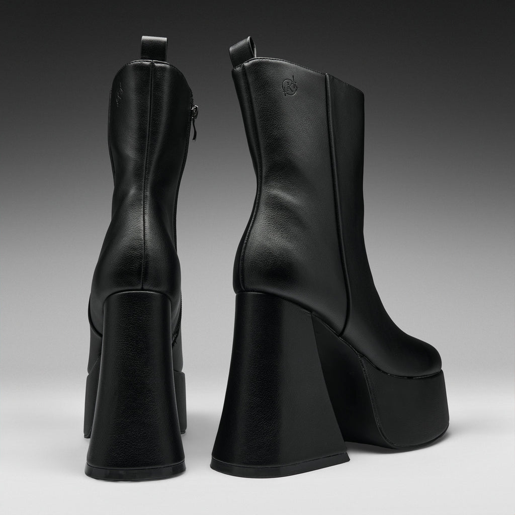 A pair of black leather ankle boots with chunky platform heels, shown from the back and side against a dark gray gradient studio background.