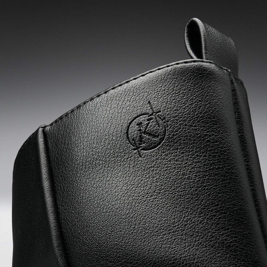 Close-up of a black textured leather bag or pouch with an embossed circular logo featuring a stylized letter K, set against a dark gray gradient background.