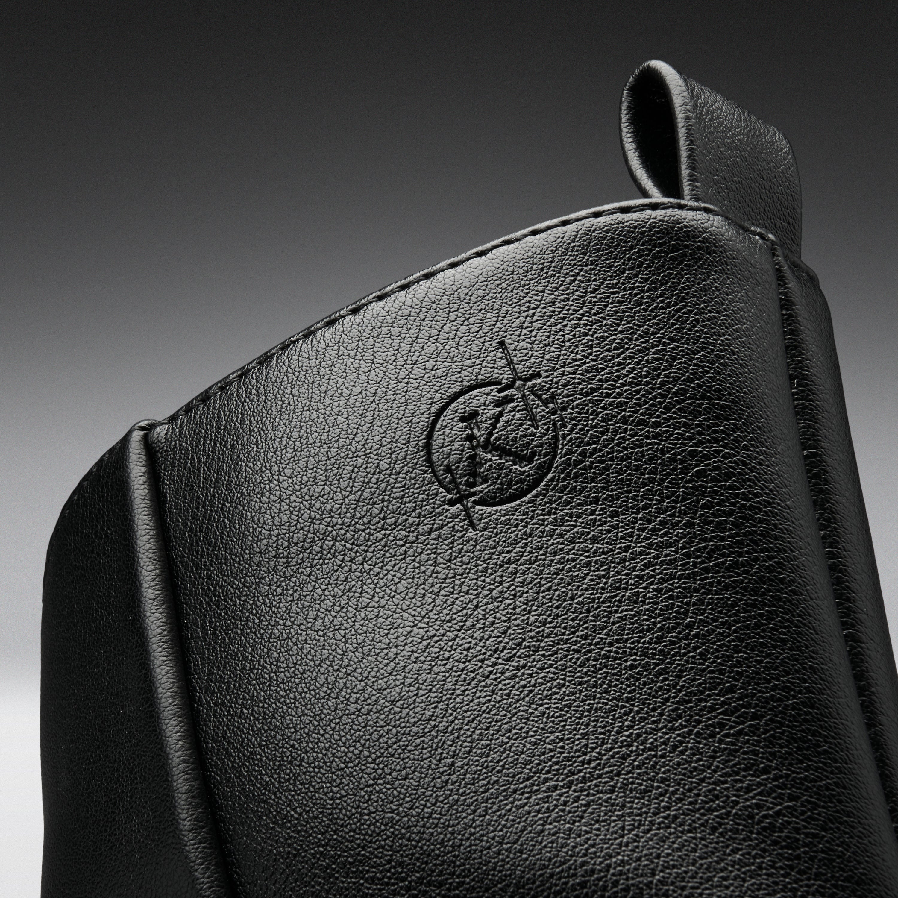 Close-up of a black textured leather bag or pouch with an embossed circular logo featuring a stylized letter K, set against a dark gray gradient background.