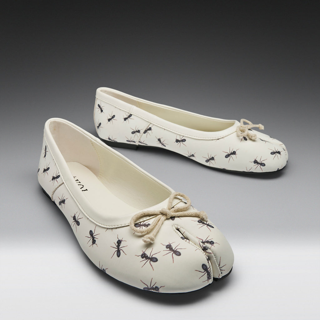 A pair of off-white split-toe ballet flats with a black ant print pattern and a beige rope bow on the toe, shown on a gray gradient background.
