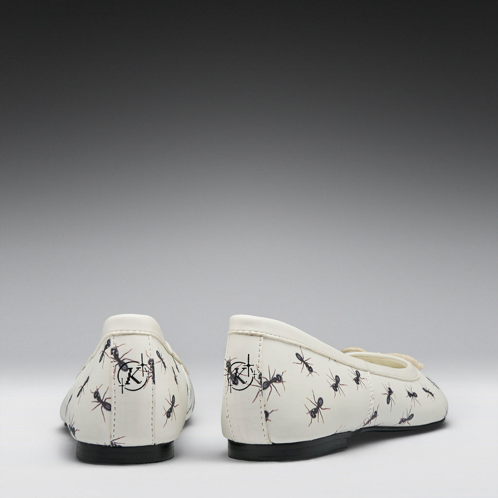 A pair of off-white ballet flats with a black ant print pattern, shown from the back against a dark-to-light gray gradient background.