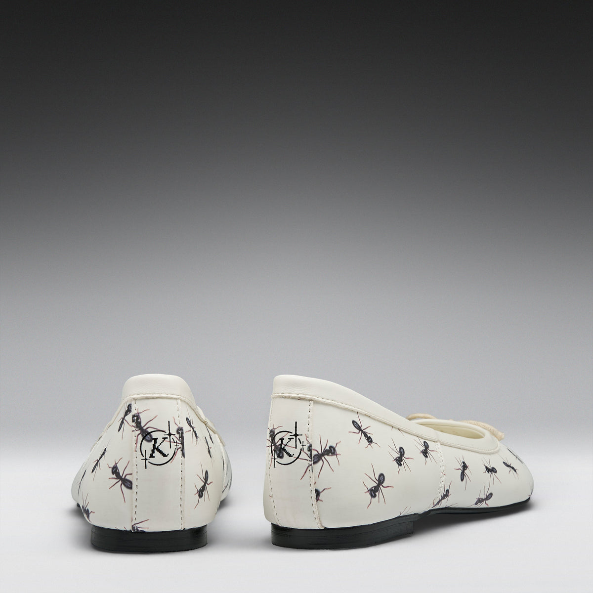 A pair of off-white ballet flats with a black ant print pattern, shown from the back against a dark-to-light gray gradient background.