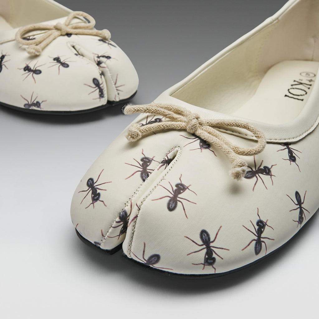 A pair of split-toe ballet flats with an off-white base, printed with black and brown ants, and finished with a beige rope bow on the top, shown on a gray gradient background.