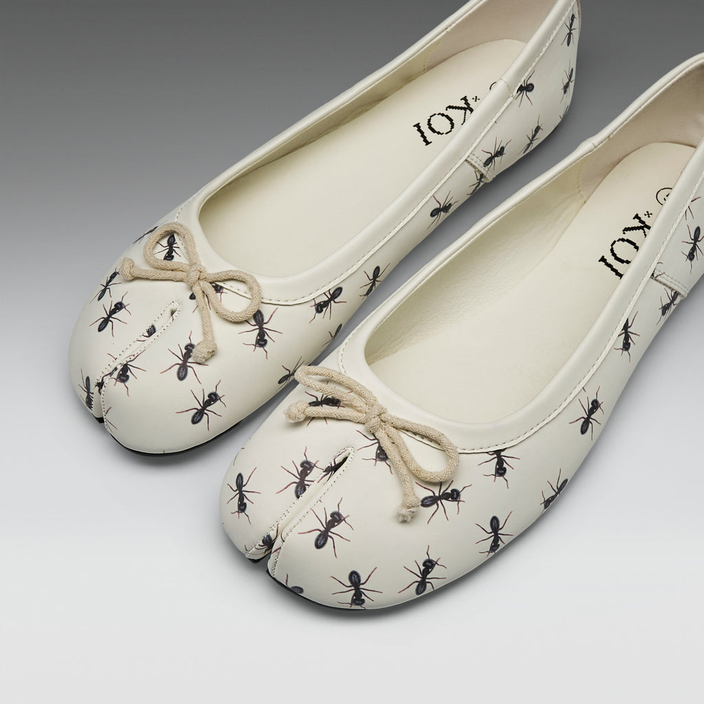 A pair of cream-colored split-toe ballet flats with a black ant print and a beige rope bow on the toe, shown on a neutral gray background.