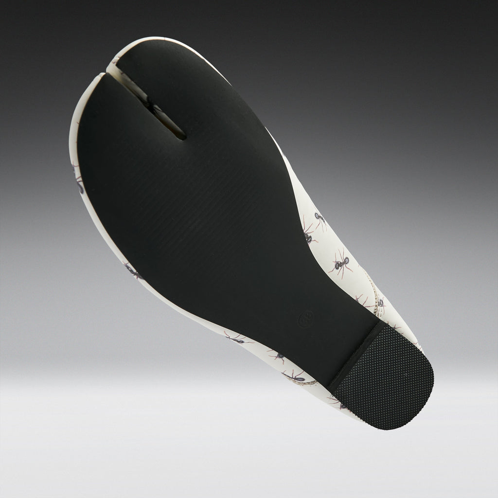 A split-foot flat sandal shown from the sole view, featuring a black textured midfoot and heel with a white and black ant-patterned outsole, set against a dark-to-light gray gradient background.