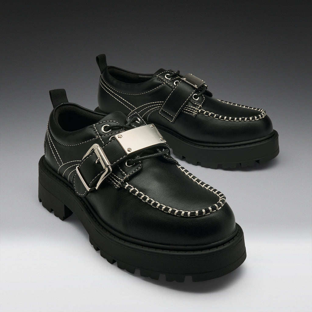 A pair of black leather platform moc-toe shoes with silver buckle straps, contrast stitching, and chunky lug soles, photographed on a dark-to-light gray gradient studio background.