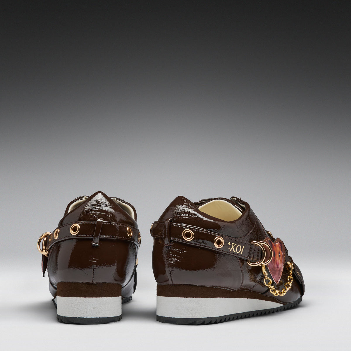 A pair of dark brown patent leather low-top sneakers shown from the back, featuring gold-tone hardware, a decorative pink and gold heart charm, and a white and black sole.