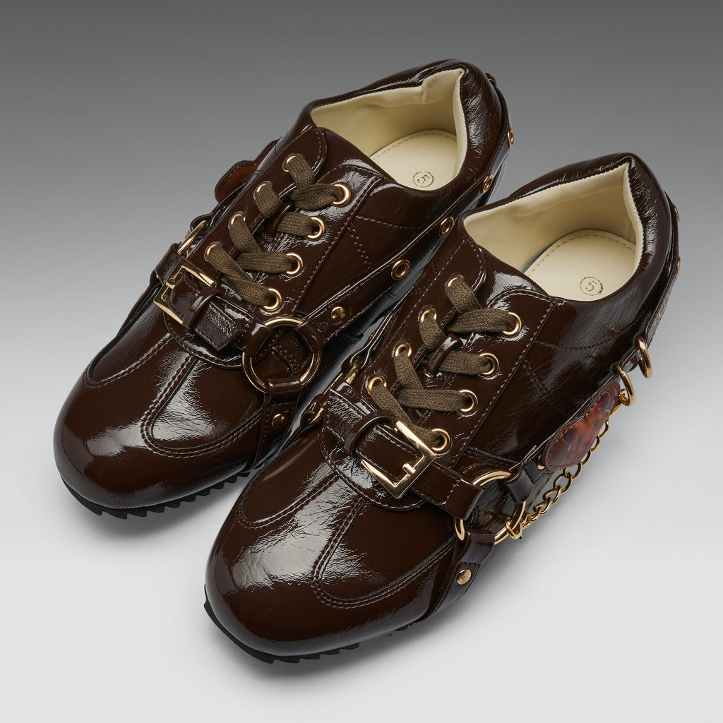 A pair of dark brown patent leather lace-up shoes with gold-tone hardware, including buckles, rings, and chain accents, shown on a neutral gray gradient background.