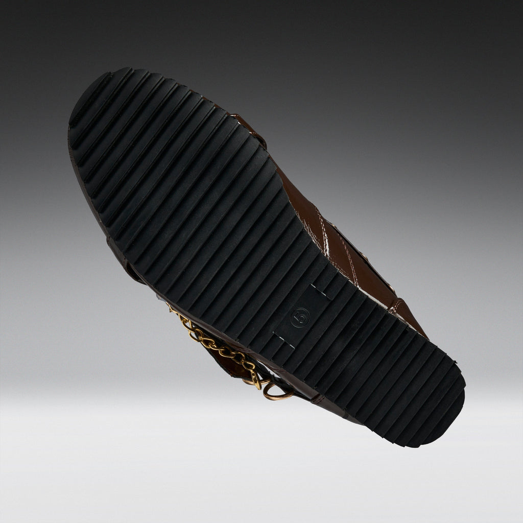 A close-up of a brown leather boot sole, showing a black ribbed rubber outsole with a small gold chain detail, set against a dark-to-light gray gradient background.