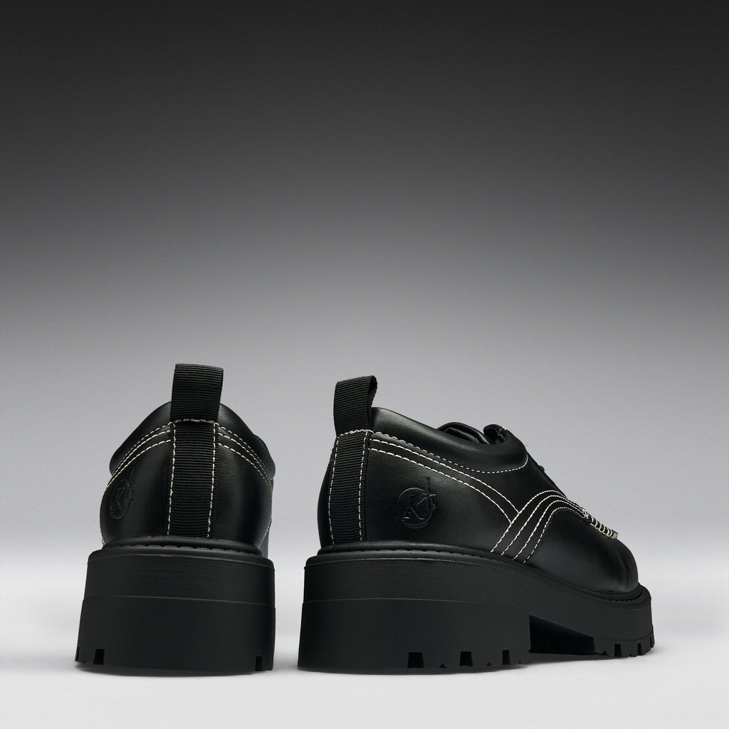 A pair of chunky black platform ankle boots with dark iridescent panels, side buckle straps, and a small circular logo on the heel, shown from the back and side against a dark gray gradient background.
