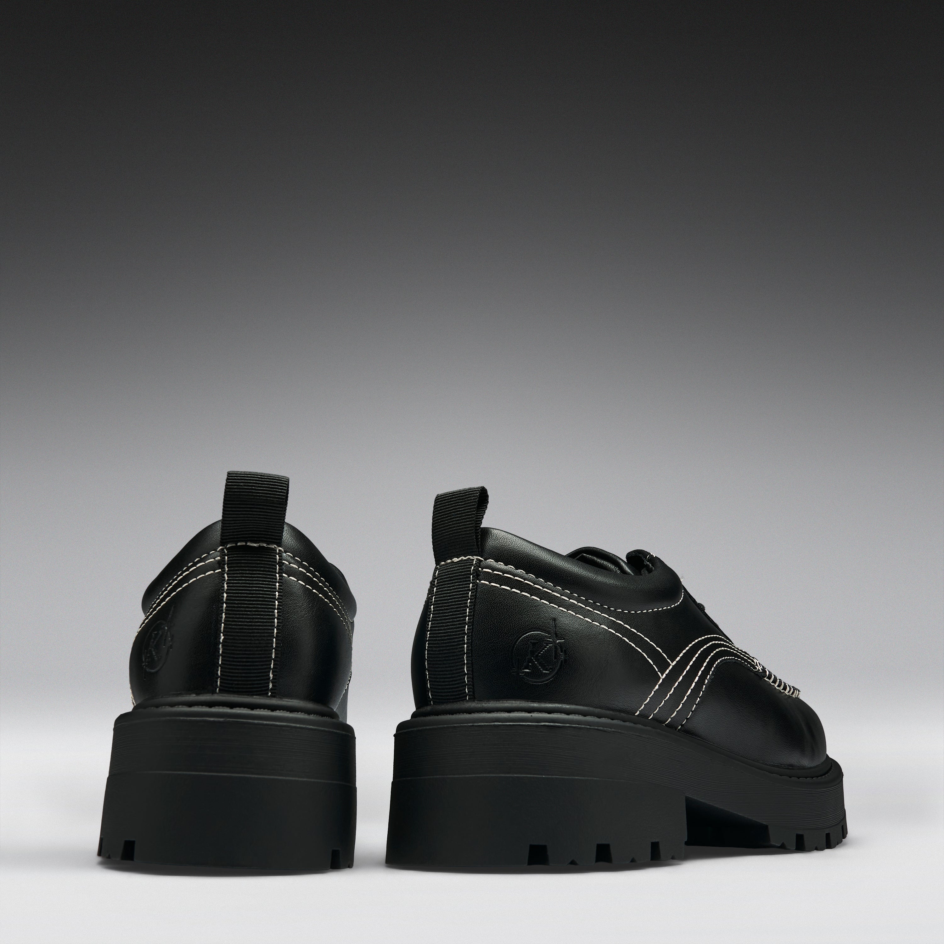 A pair of chunky black platform ankle boots with dark iridescent panels, side buckle straps, and a small circular logo on the heel, shown from the back and side against a dark gray gradient background.