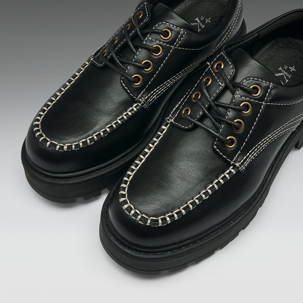 A pair of black leather lace-up shoes with distinctive white contrast stitching along the toe, gold-tone metal eyelets, and thick black soles, shown on a light gray gradient background.